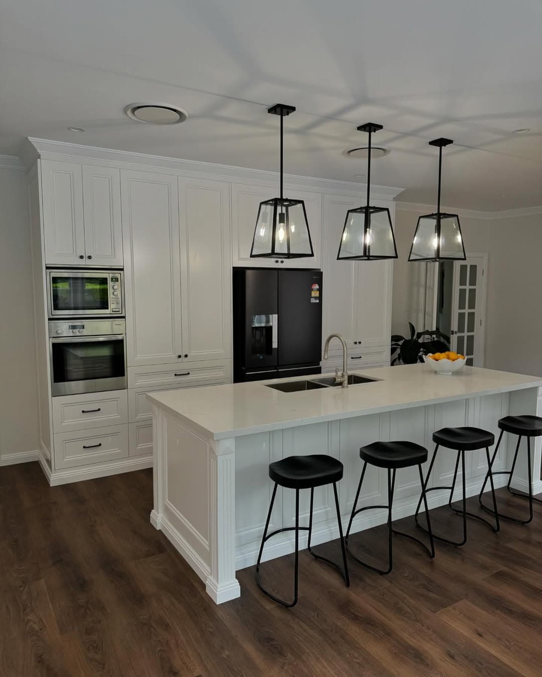 Modern white kitchen with a large island, black stools, pendant lights, and dark wood flooring.