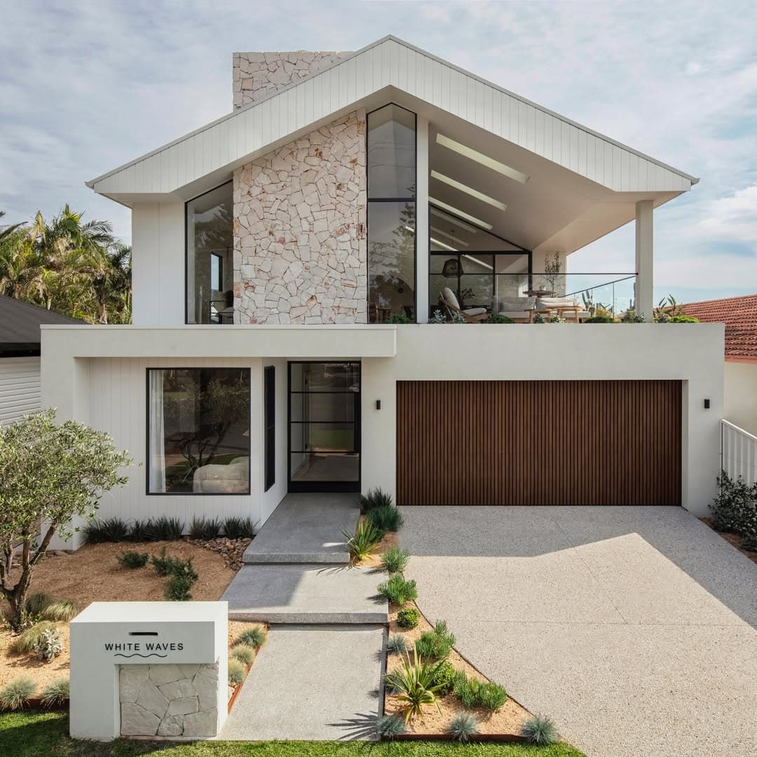 Modern two-story white house with stone accents, gabled roof, glass balcony, and wooden garage door.