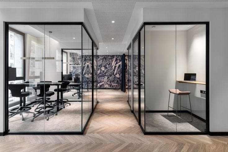 Modern office hallway with glass-walled meeting rooms, herringbone wood floors, and patterned wallpaper background.