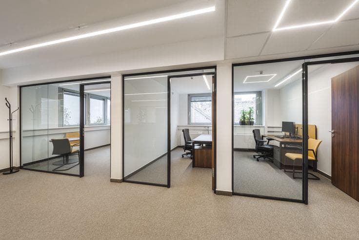 Modern office interior with glass-walled private offices, desks, chairs, and bright linear ceiling lighting.
